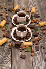 Obraz na płótnie Canvas two small white cups of coffee with, cookies, cocoa beans, slices of chocolate, hazelnuts and green leaves on wooden background