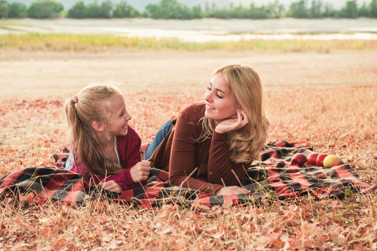Close up portrait of two smiling sisters lying under tree on a plaid. Young mom and cute daughter have a good time outsaide. Autumn weather, falling leaves.