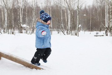 Fototapeta na wymiar Happy little boy runs on log in park during snowfall at winter d