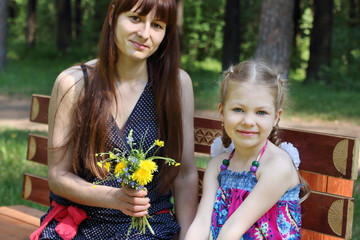 Happy mother and daughter sit with flowers on bench in park at s