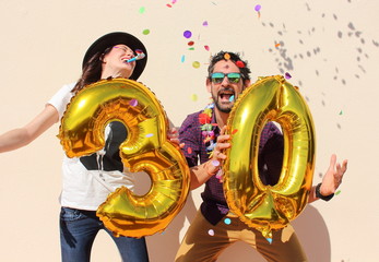Cheerful couple celebrates a thirty years birthday with big golden balloons and colorful little pieces of paper in the air.