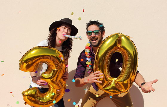 Cheerful couple celebrates a thirty years birthday with big golden balloons and colorful little pieces of paper in the air.