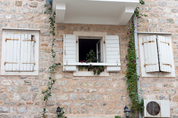 Fototapeta na wymiar Open Window with flowers and two closed windows with white shutters on a stone wall
