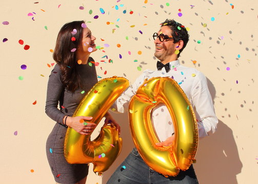 Cheerful couple celebrates a forty years birthday with big golden balloons and colorful little pieces of paper in the air.