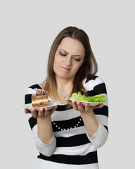 Diet, nutrition, weight loss and choice concept - Young woman chooses between cake and green lettuce on gray background