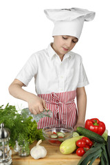 Teenage boy in cook chef hat and apron adds condiment to salad bowl on cutting board surrounded by vegetables and spices isolated on white background