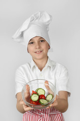 Teenage boy in cook chef hat and apron holds bowl of salad in hands with focus on bowl on gray background - vegetarian cuisine
