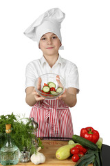Teenage boy in cook chef hat and apron holds bowl of salad in hands above table with vegetables and condiments isolated on white background - vegetarian cuisine