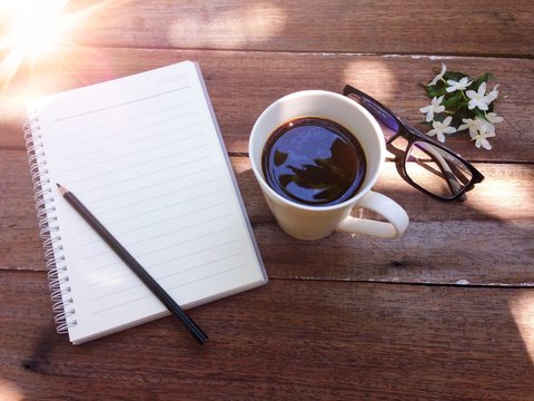 Good morning or working concept : Cup of coffee, glasses, notebook paper, pencil and white flowers on wood background and light flare