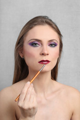 Pretty young woman with bare shoulders and bright evening makeup applies lipstick using brush on gray background