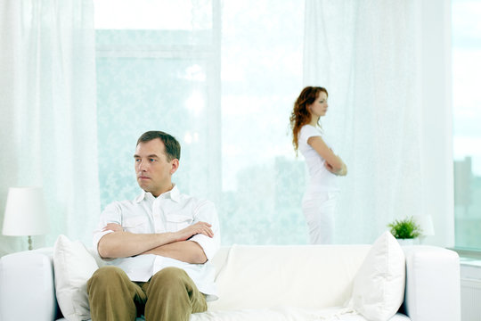 Displeased man sitting on sofa with arms crossed while his wife standing near the window