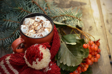 Cocoa or cup of coffee with marshmallows, fir and bright rowan branches, knitted scarf on old wooden table outdoor with beautiful bokeh lights.