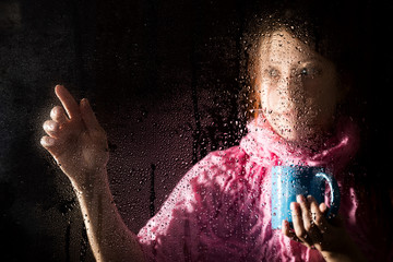 young sad woman portrait behind the window in the rain with rain drops on it. girl holding a cup of hot drink