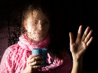 young sad woman portrait behind the window in the rain with rain drops on it. girl holding a cup of hot drink