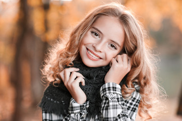 Autumn portrait of smiling blonde teen girl 13-14 year old wearing knitted scarf posing over yellow leaves outdoors. Looking at camera. Teenager hood.