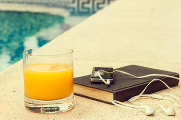 Vaso de zumo de naranja junto a un libro y reproductor de música junto a una piscina al aire libre. Vista de frente