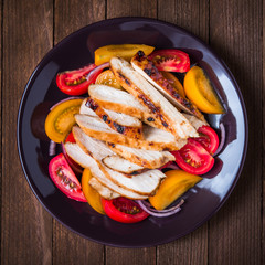 Sliced roasted chicken breast and fresh salad with tomato and purple onion top view on dark wood background. Healthy food.