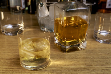Whiskey glass with ice in front of square bottle and other glasses and bottles, on wooden bar