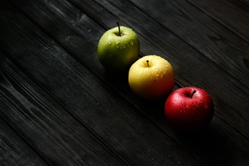 Red green yellow apples in a diagonal row with water drops on black wooden table, back light