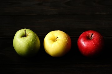 Red green yellow apples in a row with water drops on black wooden table, back light
