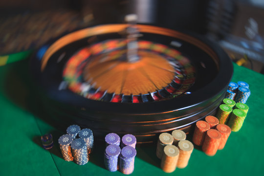A close-up vibrant image of multicolored casino table with roulette in motion, with the hand of croupier, and a group of gambling rich wealthy people in the background
