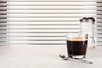 Gefüllte Kaffeetasse, Teelöffel und Zuckerstreuer auf weißer Granitplatte vor Fenster mit Jalousie