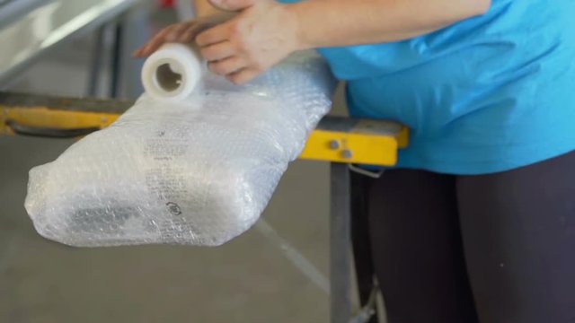 A woman in the factory is now putting a wrap around an item after it has been wrapped in a protection layer, as well. Close-up shot.
