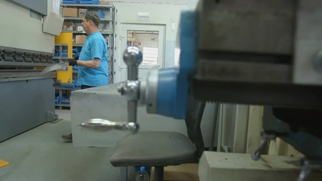 A worker in the factory is putting a long tiny metal piece under a machine in order to bend it. Wide-angle shot.
