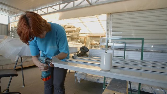 A woman in the factory is using a drilling machine to wind some screws on a metal rod. Wide-angle shot.
