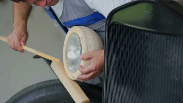 A very focused joiner is measuring the front light of the vintage car. He is making his own car. Close-up shot.

