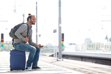 Cool man waiting at train station with mobile phone and bags