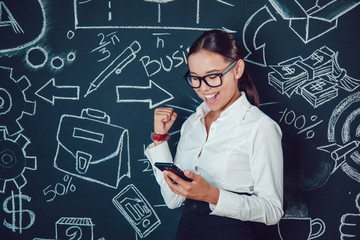 Portrait of a asian business woman in glasses standing on background with pictures and enjoys success phone in hand