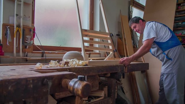 A joiner is chiselling wood in a joiner shop. He is making some nice people of wood. Wide-angle shot.
