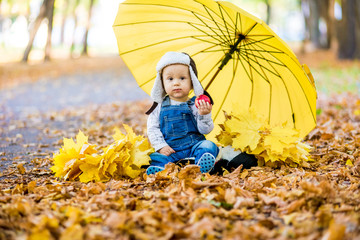 Little child sitting on grass with umbrella playing with yellow leafs in autumn day