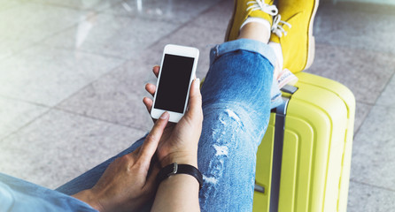 Young hipster girl at airport in yellow boot on suitcase waiting air flight, female hands holding smart phone in terminal departure lounge gate, traveler trip concept, mockup of blank screen sellphone