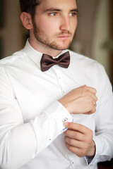 Handsome man putting on shirt standing near window at his room in morning. Preparing for some event or new workday. New opportunities, dating, wedding day or getting ready for job interview concept