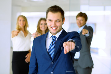 Smiling businessman  in office with colleagues in the background. Pointing by finger into the camera