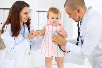 Happy cute baby  at health exam at doctor's office. Toddler girl is standing while have been keeping by two doctors