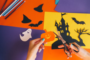 close up of female hands with scissors cut paper orange pumpkin on the background Halloween composition. Castle, ghosts, bats, pumpkins are cut from colored paper. Homemade decorations for Halloween