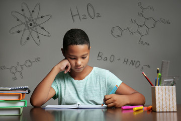 Afro-American boy studying at table. Chemical formulas on gray background. Education concept.