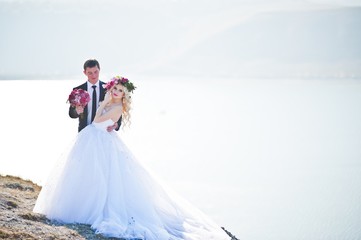 Fototapeta na wymiar Charming bride in a wreath and elegant groom on landscapes of mountains, water and blue sky at sunny weather