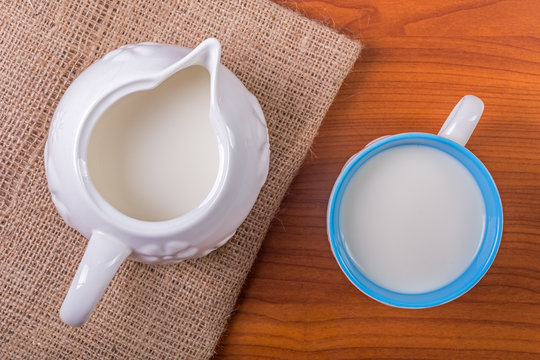 Cup and jug of milk on juta and wooden table - top view