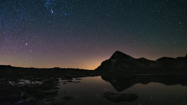 The apparent rotation of the Milky Way and the starry sky beyond snowcapped mountain ridge, reflected on idyllic apine lake. Time Lapse 4k video.