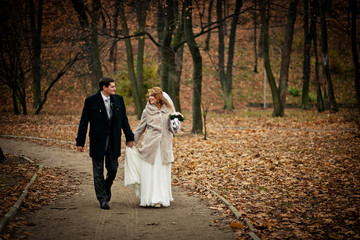 Groom holds bride's dress while they walk around autumn park