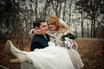 Groom carries pretty bride on his arms around autumn park
