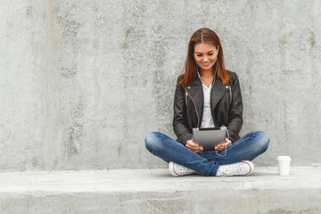 Girl with a tablet and headphones in hands sits on a concrete wall. Online education students