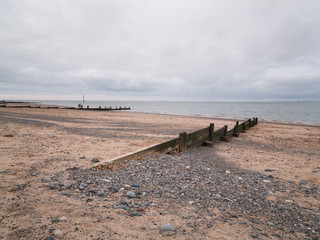 A depressing overcast gloomy day,  on a sandy beach, with weathered wooden wave breakers leading down to the sea.