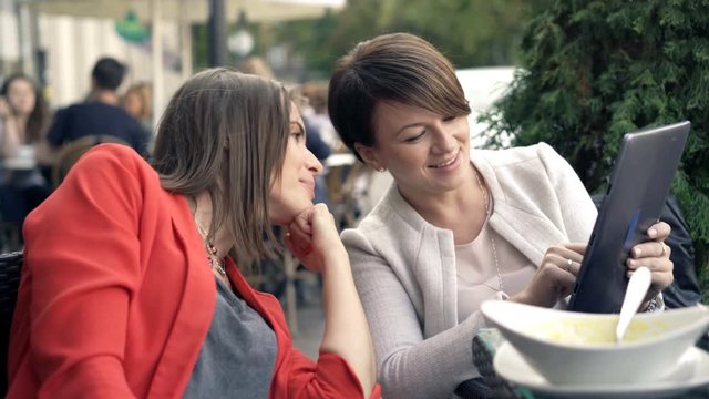 Two girlfriends talking and over tablet sitting in cafe in city
