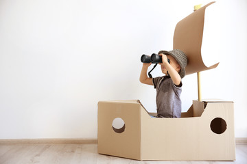 Little boy playing with cardboard ship on white wall background - obrazy, fototapety, plakaty