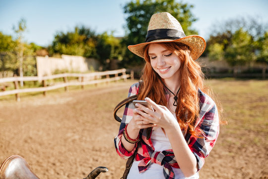 Smiling cowgirl using mobile phone while standing at ranch fence